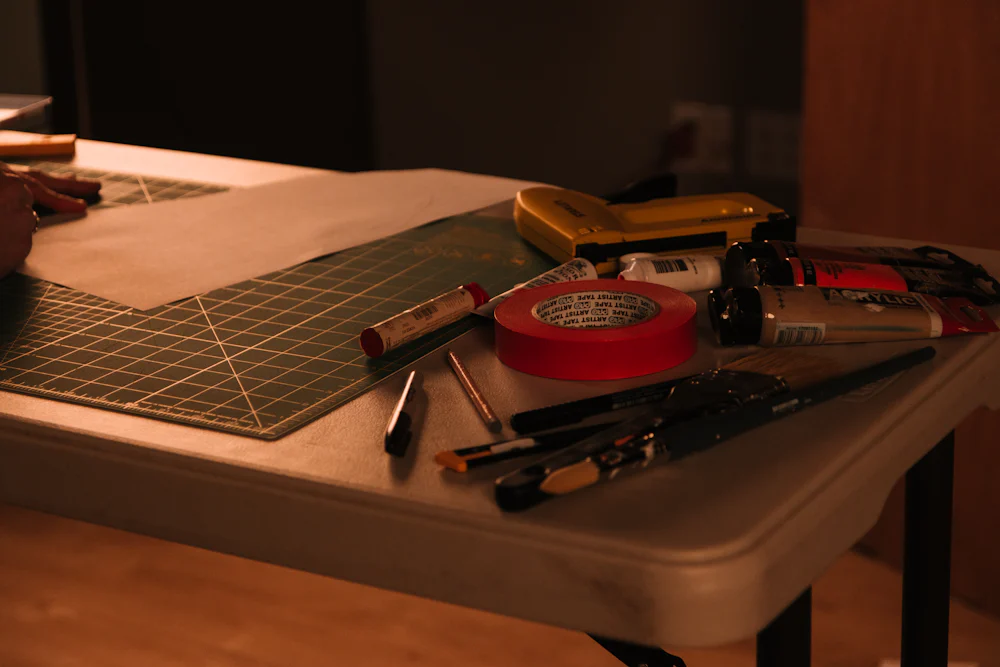 A workbench with a cutting mat, tape, a ruler and cutting tools under warm light